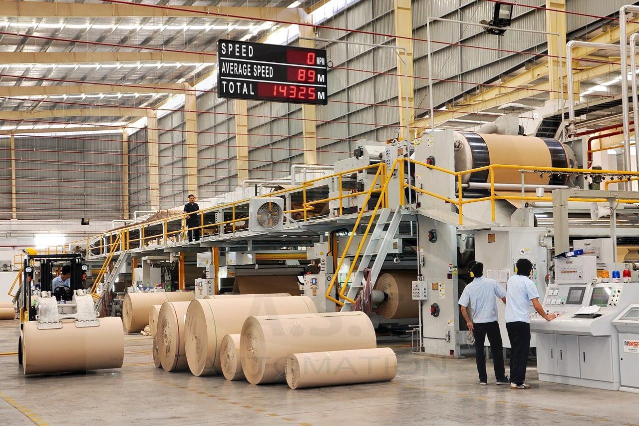 Hình ảnh dây chuyền sản xuất carton trong nhà máy với các cuộn giấy nguyên liệu và nhiều cụm thiết bị hoạt động liên tục, yêu cầu phối hợp tốc độ giữa các công đoạn.