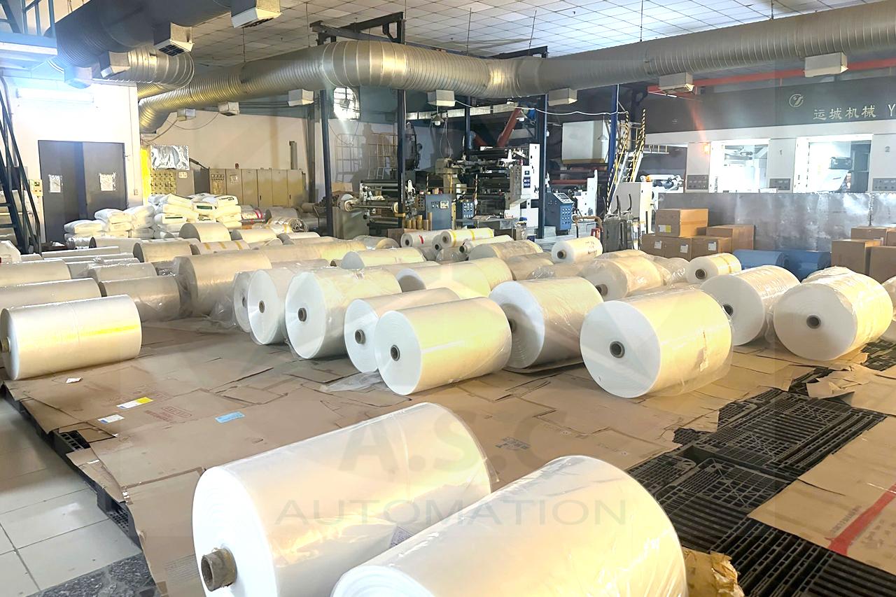 Các cuộn vật liệu dạng màng trong khu vực sản xuất bao bì, phía sau là hệ thống máy xử lý vật liệu.
